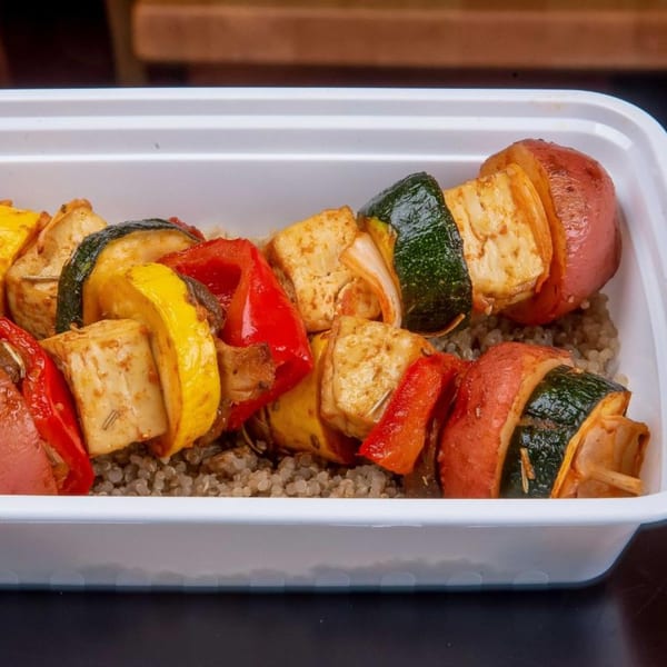 Two colorful vegetable and tofu skewers over quinoa in a white takeout container.