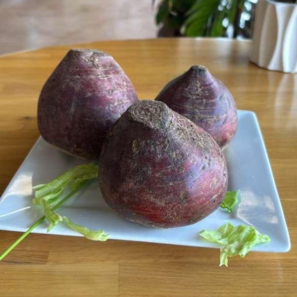 Three dark red beets with green stems on a white rectangular plate on a wooden table.