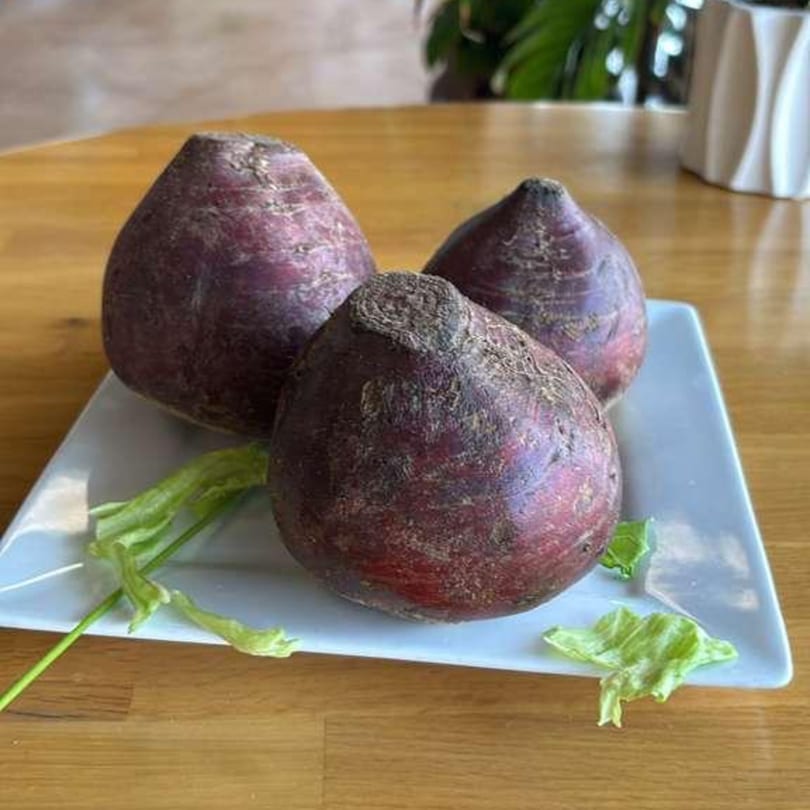 Three dark red beets with green stems on a white rectangular plate on a wooden table.
