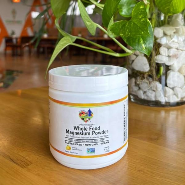 White container of "Whole Food Magnesium Powder" on a wooden table with a plant in the background.