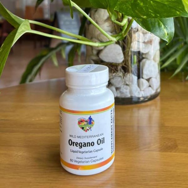 A white bottle of Wild Mediterranean Oregano Oil capsules sits on a wooden table next to a plant.