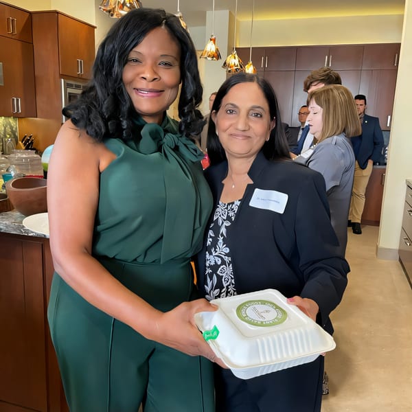Two women smiling, one in green, the other in black, holding a FoodAMed takeout container.