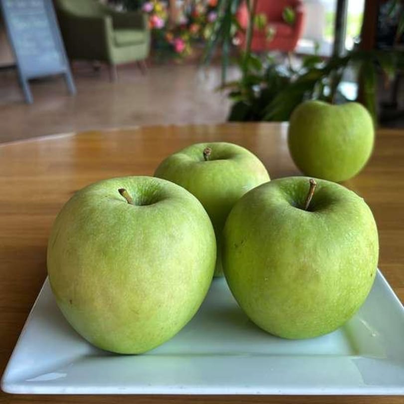 Four green apples on a white rectangular plate, with a blurred restaurant interior in the background.