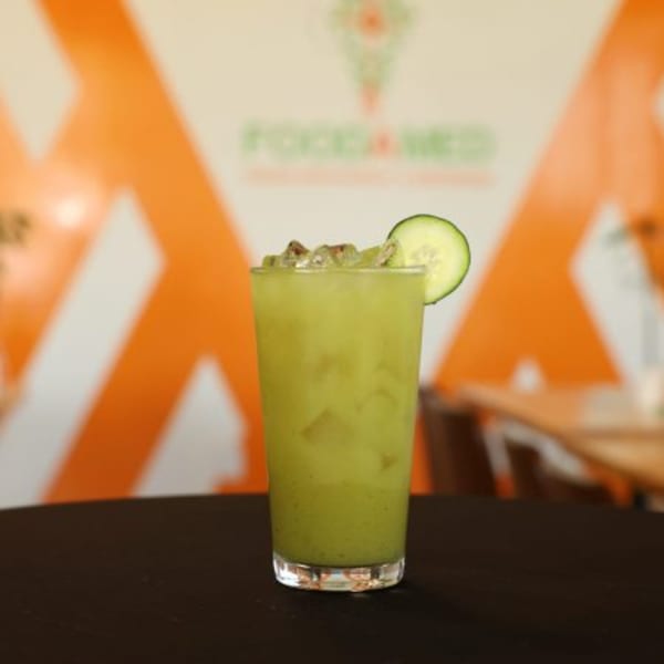 A tall glass of green juice with ice and a cucumber slice, on a dark table at FoodAMed.
