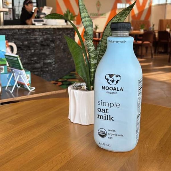 Mooala organic oat milk bottle on a wooden table with a plant and a blurred restaurant background.