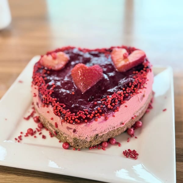 Heart-shaped pink cheesecake with berry topping and red sprinkles on a white plate.
