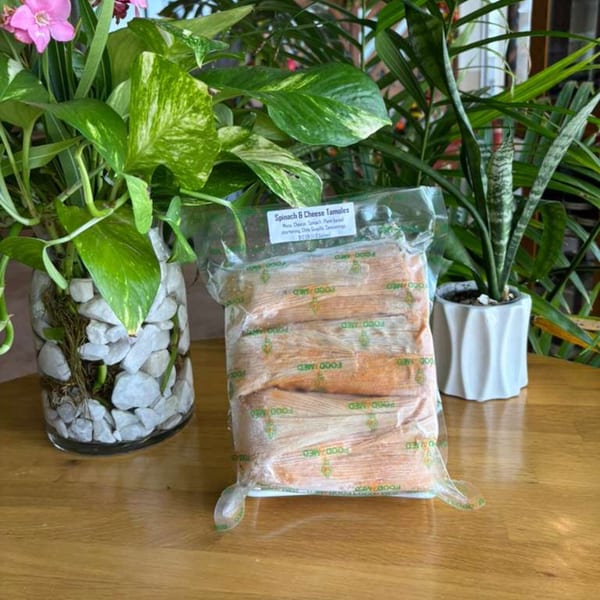 A vacuum-sealed package of spinach and cheese tamales sits on a wooden table surrounded by potted plants.