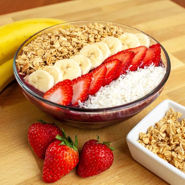 A FoodAMed acai bowl with granola, banana slices, strawberries, and shredded coconut on a wooden table.