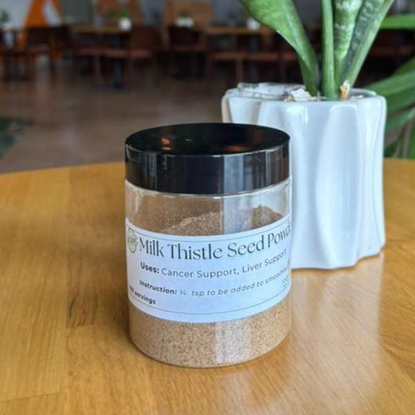 A clear jar of Milk Thistle Seed Powder sits on a wooden table next to a potted plant.