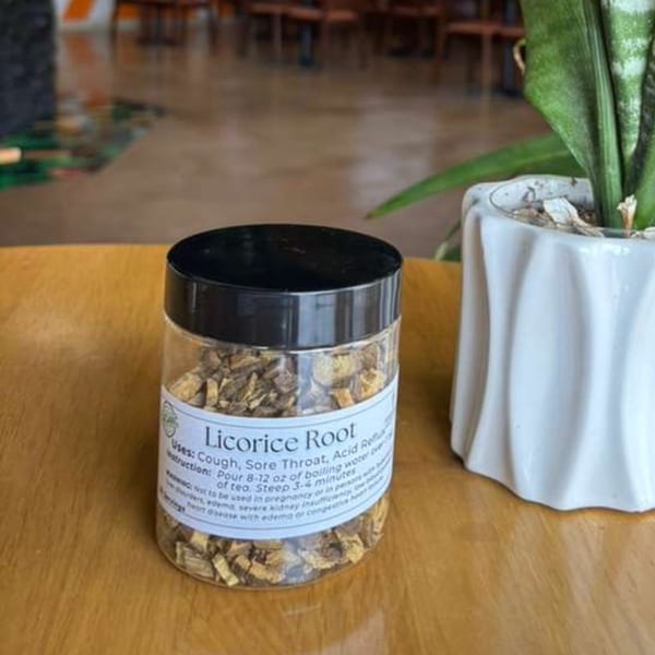 A clear jar of licorice root pieces with a black lid sits on a wooden table next to a potted plant.