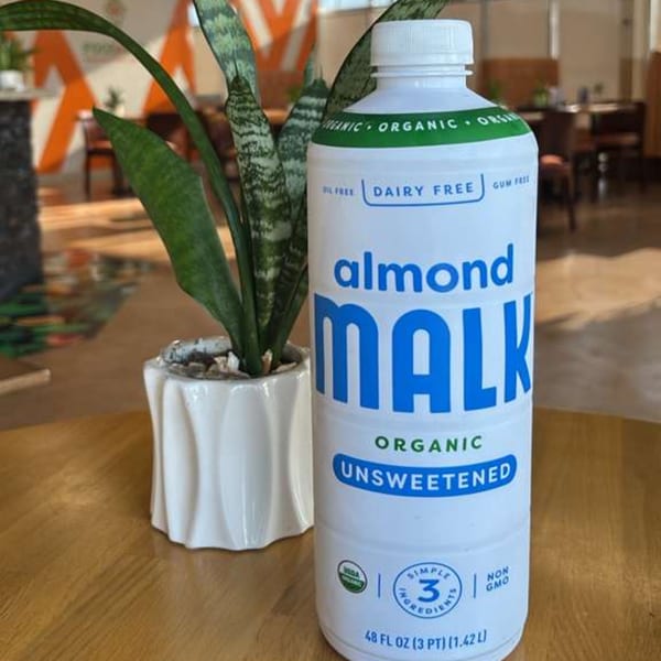 A bottle of Malk almond milk sits on a wooden table next to a potted snake plant.