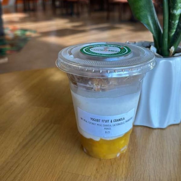 A Foodamed yogurt fruit and granola cup with mango on a wooden table next to a plant.