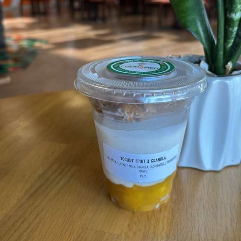 A Foodamed yogurt fruit and granola cup with mango on a wooden table next to a plant.