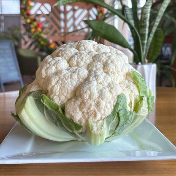 A whole head of fresh cauliflower with green leaves rests on a white square plate.