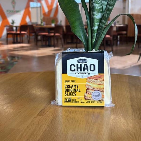 Chao Creamery dairy-free cheese slices on a wooden table with a plant and restaurant interior in the background.