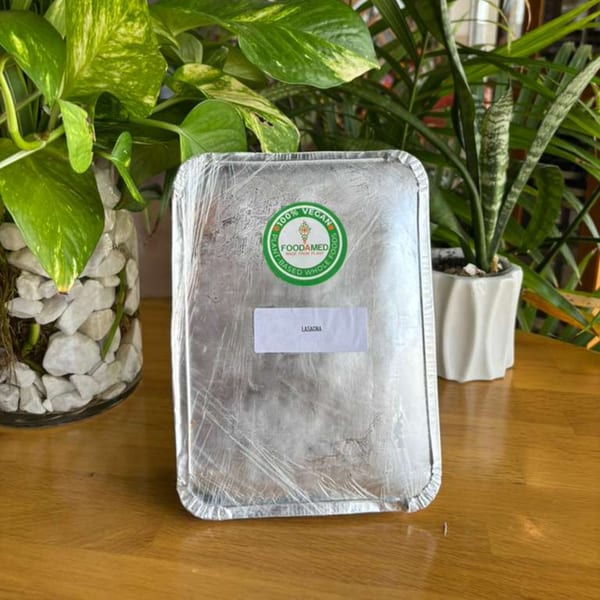 A foil container of vegan lasagna from Foodamed sits on a wooden table, surrounded by green plants.