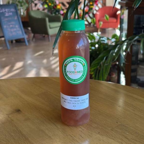 A bottle of Foodamed Kombucha with a green cap sits on a wooden table in a restaurant.